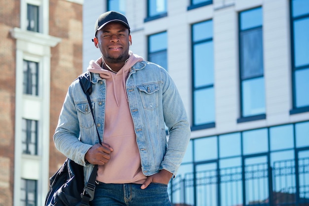 Conceito de moda de rua elegante homem afro-americano bonito em um capuz de camisa rosa e jaqueta jeans e boné preto contra as janelas do centro de negócios