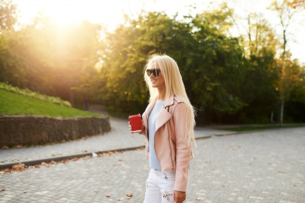 Conceito de moda de rua. estudante jovem elegante andando no parque ensolarado e detém uma xícara de café para ir.