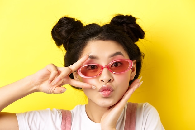 Conceito de moda de verão. Linda e glamourosa garota asiática de óculos escuros, mostrando o sinal de v kawaii e olhando para a câmera, em pé sobre fundo amarelo