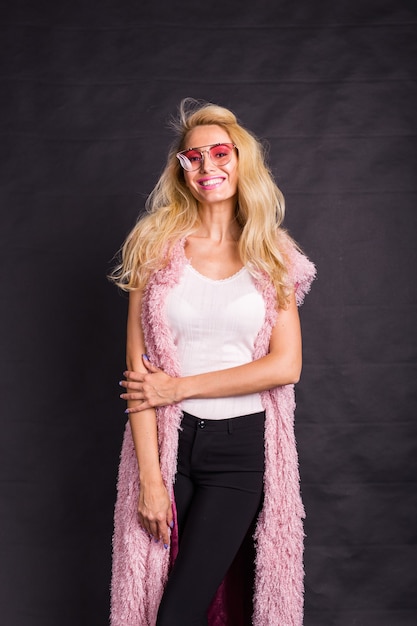 Conceito de moda e beleza - modelo loira com óculos cor de rosa, vestida com camisa branca e casaco de lã rosa sobre a superfície escura.