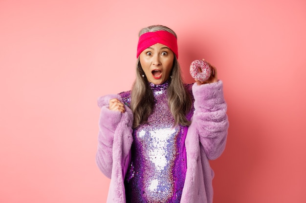 Conceito de moda e comida. Linda mulher asiática sênior em um vestido elegante e brilhante segurando uma rosquinha doce e gritando para a câmera, com medo de calorias, fundo rosa