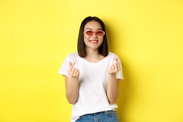 Conceito de moda e estilo de vida. Mulher asiática atraente em elegantes óculos de sol, mostrando os corações dos dedos e sorrindo feliz para a câmera, em pé sobre amarelo.
