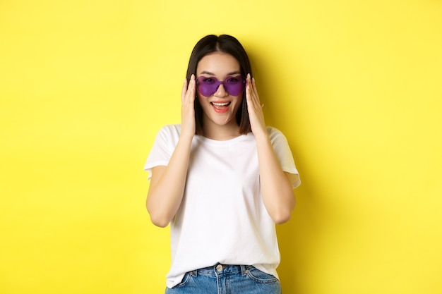 Conceito de moda e estilo de vida. Mulher asiática elegante experimentando novos óculos de sol em formato de coração, saindo de férias, sorrindo feliz para a câmera, em pé sobre fundo amarelo.