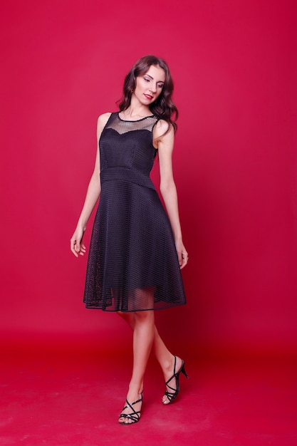 Conceito de moda, estilo e pessoas - mulher jovem e bonita em um elegante vestido de cocktail preto está segurando a mão no queixo, olhando para longe e pensando. estúdio de comprimento de três quartos baleado em fundo rosa.