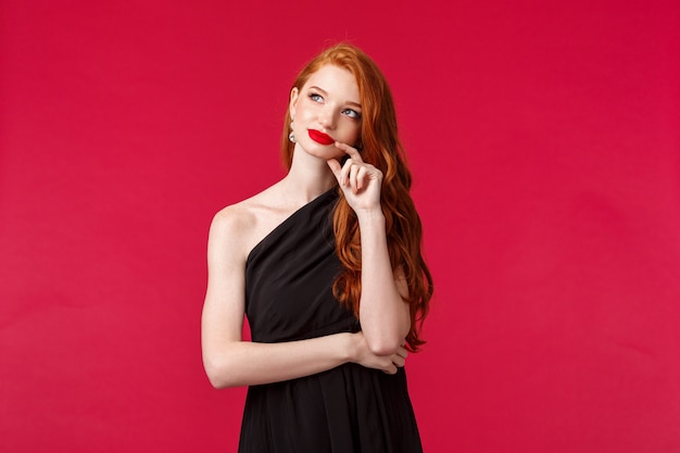 Conceito de moda, luxo e beleza. Retrato de mulher ruiva jovem elegante e sensual sonhadora com batom vermelho, maquiagem de noite, use vestido preto luxuoso, procure como sonho ou imagem