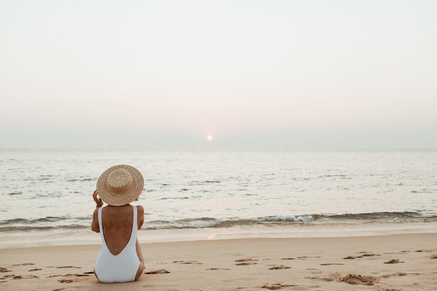 Conceito de moda para férias de verão Uma jovem bronzeada vestindo um lindo maiô branco com um chapéu de palha está sentada e relaxando em uma praia tropical com areia branca e está assistindo o pôr do sol e o mar