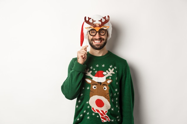 Conceito de Natal e feriados. Homem de urso sorridente com chapéu de Papai Noel, parecendo feliz, segurando uma máscara de festa para o ano novo, comemorando o natal, em pé sobre um fundo branco