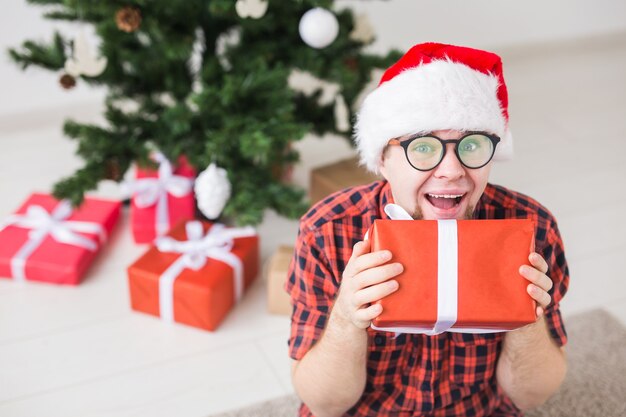 Conceito de Natal e férias - engraçado homem com chapéu de Papai Noel segurando um presente em casa na sala de estar.