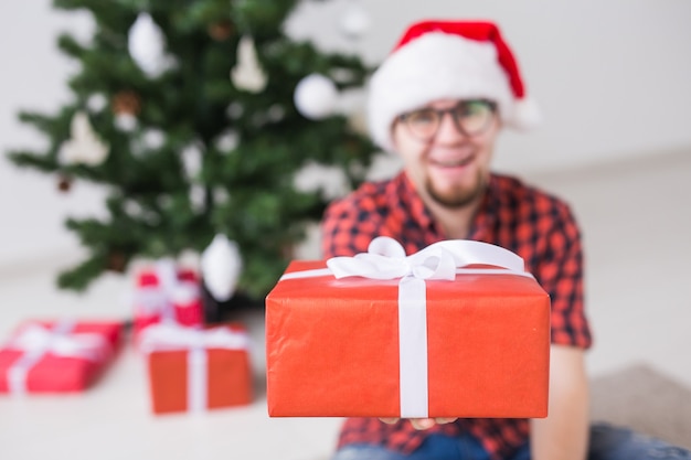 Conceito de Natal e férias - engraçado homem com chapéu de Papai Noel segurando um presente em casa na sala de estar.