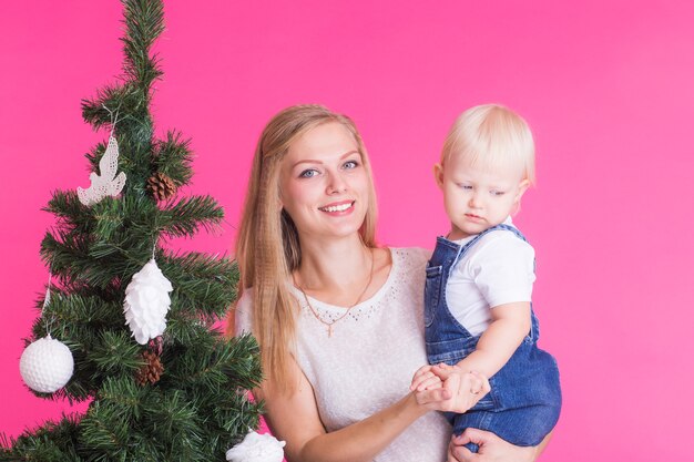 Conceito de Natal e férias - retrato de uma mulher sorridente, segurando sua filha perto de árvore de Natal na parede rosa.