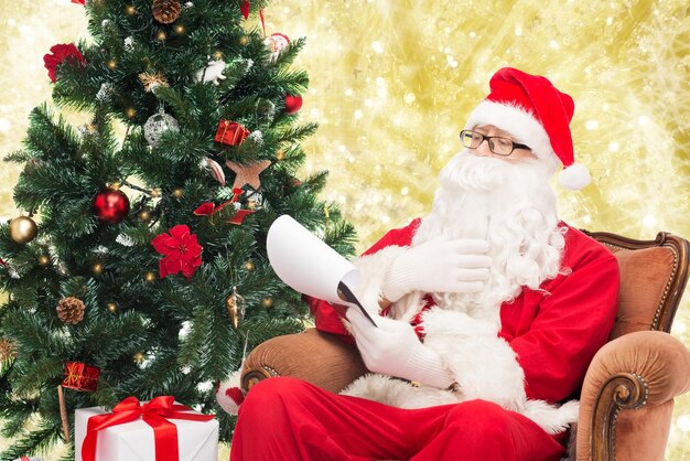 conceito de natal, feriados e pessoas - homem fantasiado de papai noel com bloco de notas e caneta sobre fundo de luzes amarelas