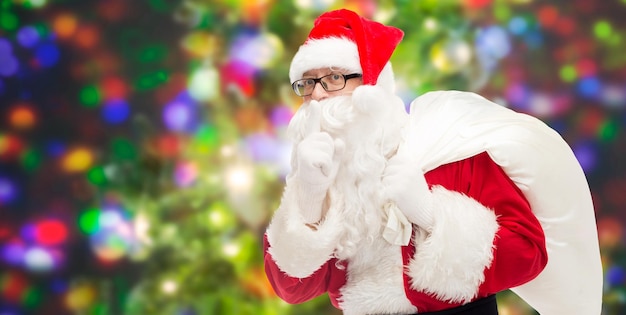 conceito de natal, feriados e pessoas - homem fantasiado de papai noel com bolsa fazendo gesto de silêncio sobre fundo de luzes de festa