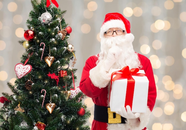 Conceito de natal, feriados e pessoas - homem fantasiado de papai noel com caixa de presente e árvore fazendo gesto de silêncio sobre fundo de luzes