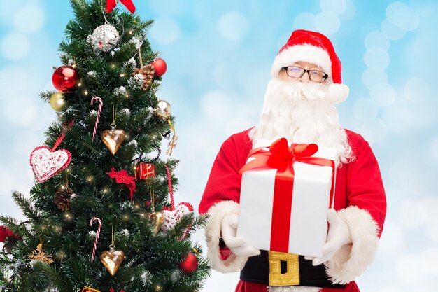 conceito de natal, feriados e pessoas - homem fantasiado de papai noel com caixa de presente e árvore sobre fundo de luzes azuis