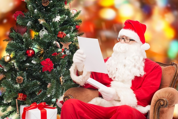 conceito de natal, feriados e pessoas - homem fantasiado de papai noel com carta e árvore de natal sentado na poltrona sobre fundo de luzes vermelhas