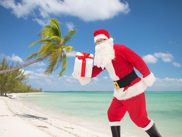 conceito de natal, feriados e pessoas - homem fantasiado de papai noel correndo com caixa de presente sobre fundo de praia tropical