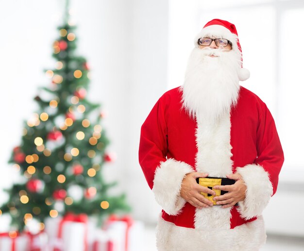 conceito de natal, feriados e pessoas - homem fantasiado de papai noel na sala de estar com fundo de árvore