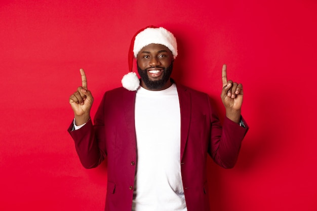 Conceito de Natal, festa e feriados. Homem afro-americano bonito com chapéu de Papai Noel sorrindo, apontando o dedo para cima e mostrando um anúncio, em pé sobre um fundo vermelho