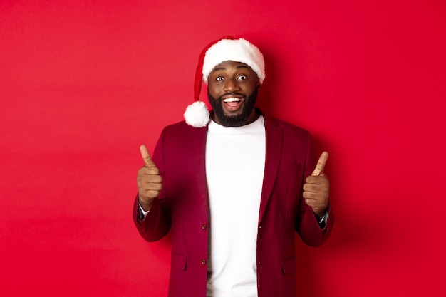 Conceito de Natal, festa e feriados. Homem negro alegre com chapéu de Papai Noel mostrando o polegar para cima, comemorando o ano novo, aprovo e concordo, em pé contra um fundo vermelho