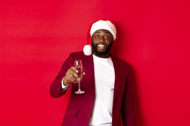 Conceito de Natal, festa e feriados. Homem negro alegre dizendo "brinde", levantando a taça de champanhe e desejando feliz ano novo, de pé contra um fundo vermelho.