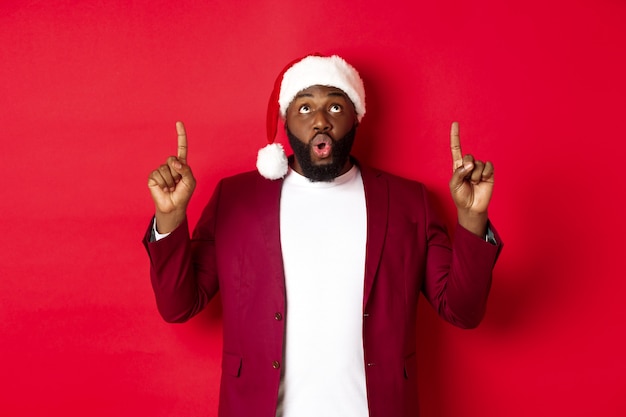 Conceito de Natal, festa e feriados. Homem negro impressionado com chapéu de Papai Noel apontando o dedo para cima, dizendo uau e olhando para a câmera, verificando a oferta promocional, fundo vermelho