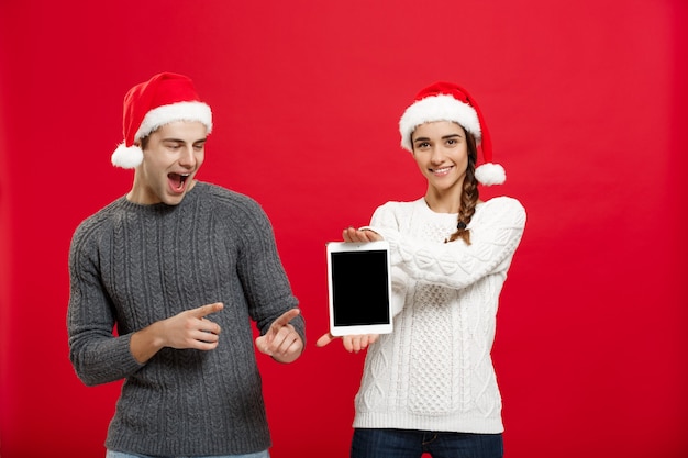 Conceito de natal - jovem casal feliz em suéteres de natal surpreendente algo no tablet digital.