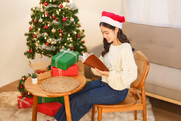 Conceito de Natal Jovem mulher asiática sentada na cadeira perto da árvore de natal e lendo um livro