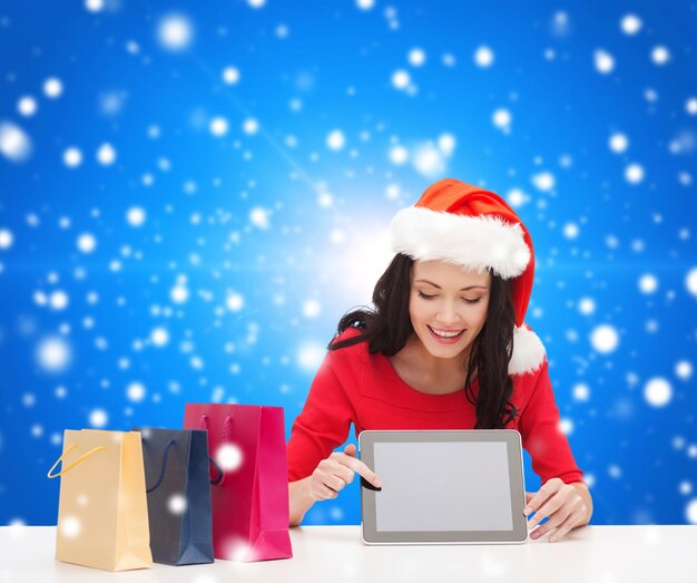 conceito de natal, tecnologia e pessoas - mulher sorridente no chapéu de ajudante de papai noel com sacolas de compras e computador tablet pc sobre fundo azul nevado