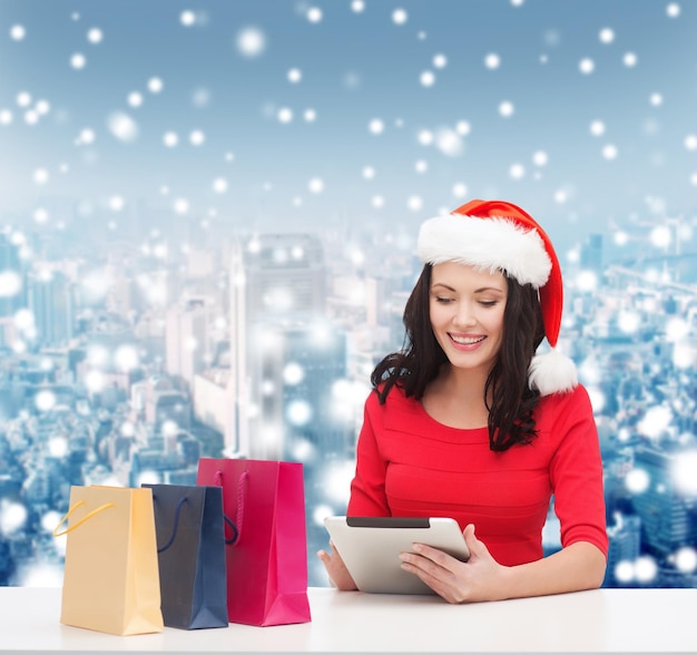 conceito de natal, tecnologia e pessoas - mulher sorridente no chapéu de ajudante de papai noel com sacolas de compras e computador tablet pc sobre o fundo da cidade de neve