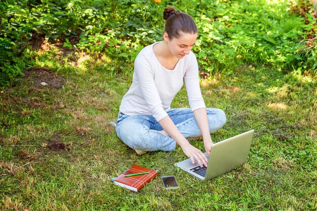 Conceito de negócio freelance. Jovem mulher sentada no gramado verde no parque da cidade trabalhando no computador portátil. Garota de estudante sincera autêntica de estilo de vida estudando ao ar livre. Escritório Móvel