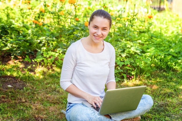 Conceito de negócio freelance. Jovem mulher sentada no gramado verde no parque da cidade trabalhando no computador portátil. Garota de estudante sincera autêntica de estilo de vida estudando ao ar livre. Escritório Móvel