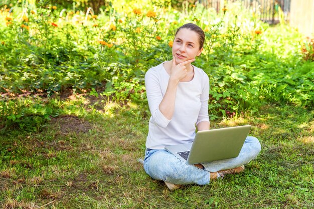 Conceito de negócio freelance. Jovem mulher sentada no gramado verde no parque da cidade trabalhando no computador portátil. Garota de estudante sincera autêntica de estilo de vida estudando ao ar livre. Escritório Móvel