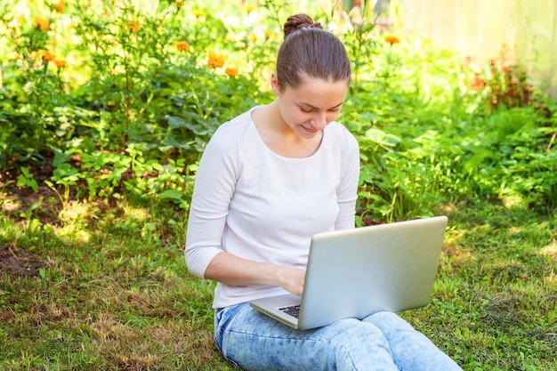 Conceito de negócio freelance. Jovem mulher sentada no gramado verde no parque da cidade trabalhando no computador portátil. Garota de estudante sincera autêntica de estilo de vida estudando ao ar livre. Escritório Móvel