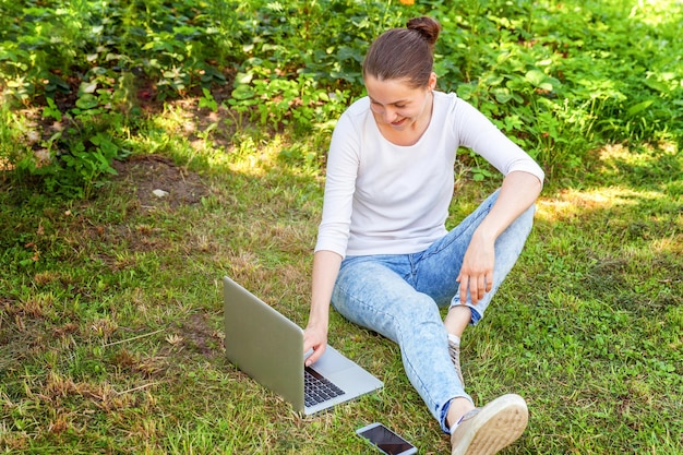 Conceito de negócio freelance. Jovem mulher sentada no gramado verde no parque da cidade trabalhando no computador portátil. Garota de estudante sincera autêntica de estilo de vida estudando ao ar livre. Escritório Móvel