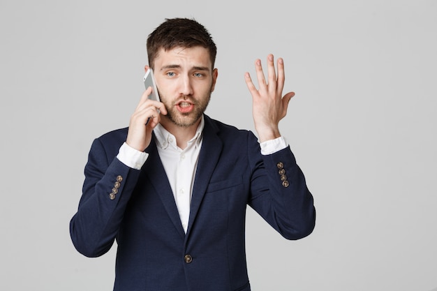 Conceito de negócio - Retrato jovem e bonito homem de negócios irritado em terno falando no telefone olhando para a câmera. Fundo branco.