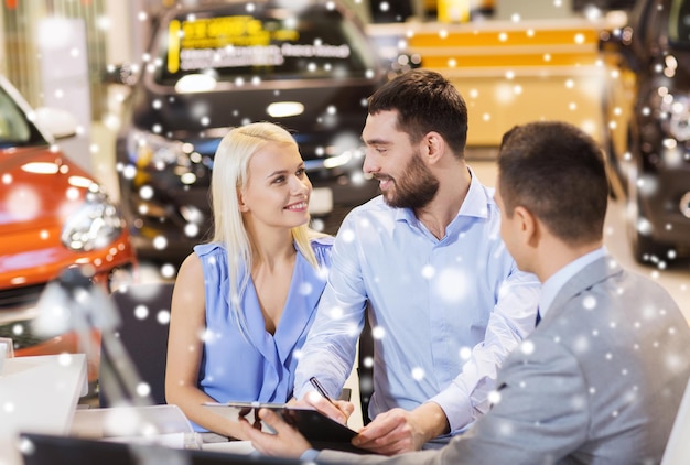 Conceito de negócio, venda e pessoas de automóveis - casal feliz com revendedor comprando carro em salão de automóveis ou salão sobre efeito de neve