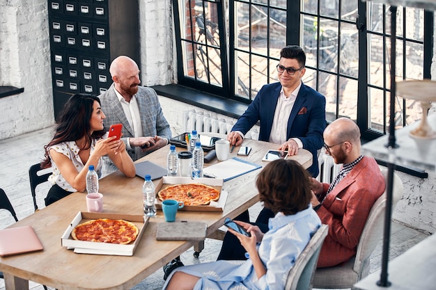 Conceito de negócios, comida, almoço e pessoas - equipe de negócios internacionais feliz comendo pizza no escritório.