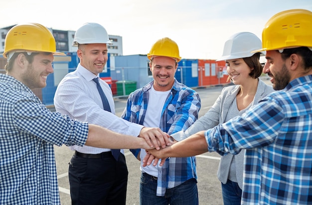 Conceito de negócios, construção, parceria, gesto e pessoas - sorridentes construtores e arquitetos em capacetes com as mãos no topo ao ar livre