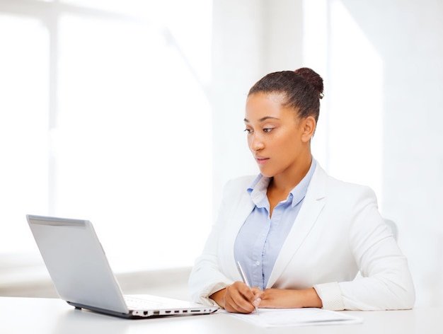 conceito de negócios e tecnologia - empresária africana com laptop escrevendo algo no caderno