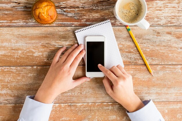 conceito de negócios, educação, pessoas e tecnologia - close-up de mãos femininas tocando a tela em branco do smartphone com notebook, muffin e xícara de café na mesa