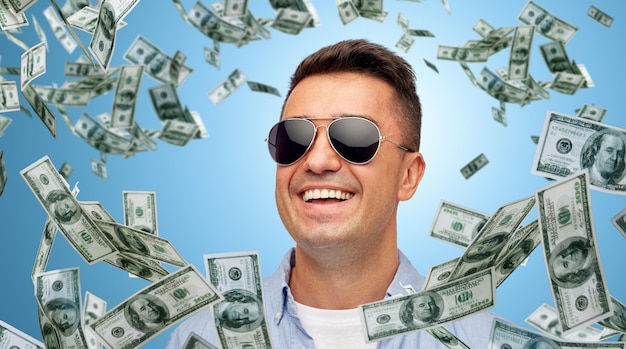 Conceito de negócios, finanças, sucesso e pessoas - rosto de homem latino de meia-idade sorridente na camisa e óculos de sol sobre fundo azul com um monte de dinheiro em dólar caindo