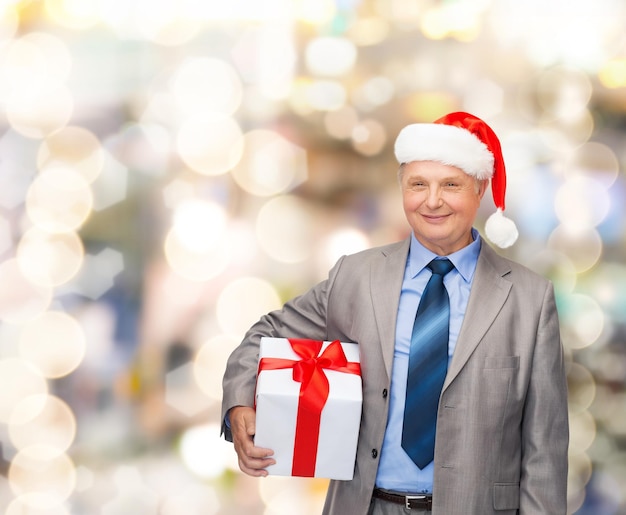 Conceito de negócios, natal, presentes e pessoas - homem sênior sorridente de terno e chapéu de ajudante de papai noel com presente sobre fundo de luzes