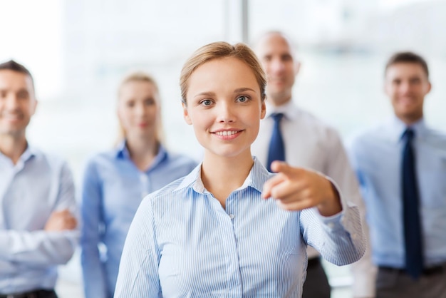 conceito de negócios, pessoas e trabalho em equipe - empresária sorridente apontando o dedo em você com grupo de empresários no escritório