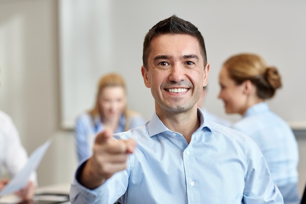 conceito de negócios, pessoas, gestos e trabalho em equipe - empresário sorridente apontando o dedo para você com grupo de empresários reunidos no escritório