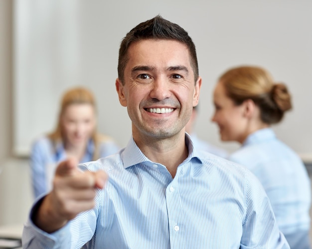 Foto conceito de negócios, pessoas, gestos e trabalho em equipe - empresário sorridente apontando o dedo para você com grupo de empresários reunidos no escritório