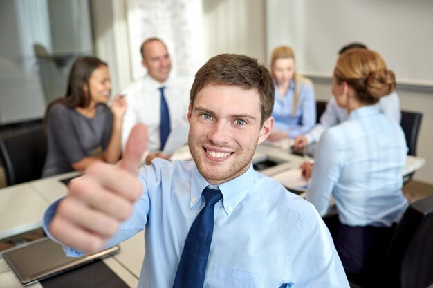 Foto conceito de negócios, pessoas, gestos e trabalho em equipe - empresário sorridente mostrando polegares para cima com grupo de empresários reunidos no escritório