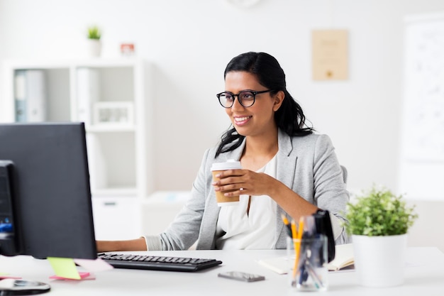 conceito de negócios, pessoas, trabalho e tecnologia - empresária feliz com computador tomando café em copo de papel no escritório