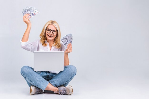 Conceito de negócios. Retrato de mulher envelhecida sênior feliz casual sentado no chão em posição de lótus e segurando o laptop e contas de dinheiro isoladas no fundo branco. Copie o espaço para o texto.