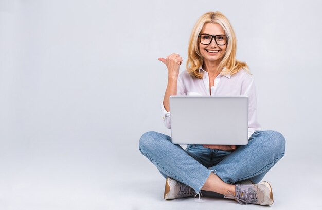 Conceito de negócios. Retrato de mulher idosa sênior feliz casual sentado no chão em posição de lótus e segurando laptop isolado no fundo branco. Copie o espaço para o texto.