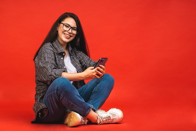 Conceito de negócios retrato de mulher morena feliz em casual sentado no chão em pose de lótus e usando telefone celular isolado sobre fundo vermelho